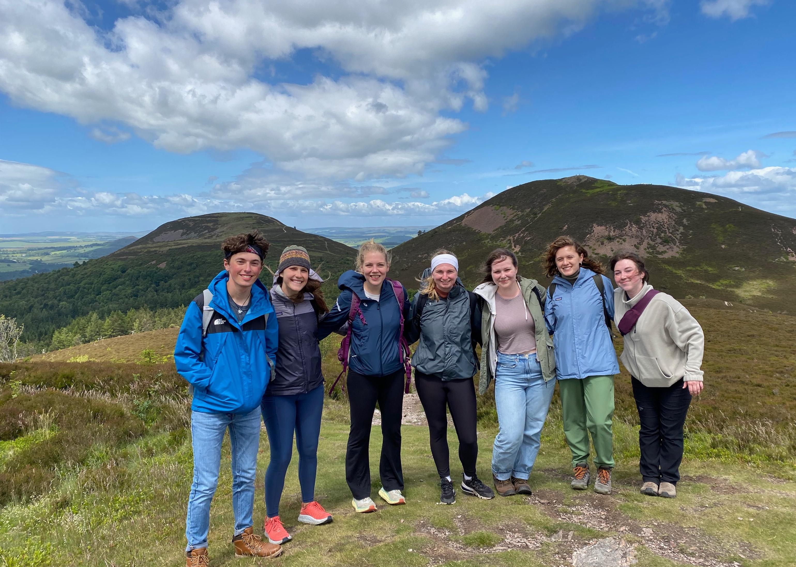 Group of seven people standing in front of a mountain on a windy day.