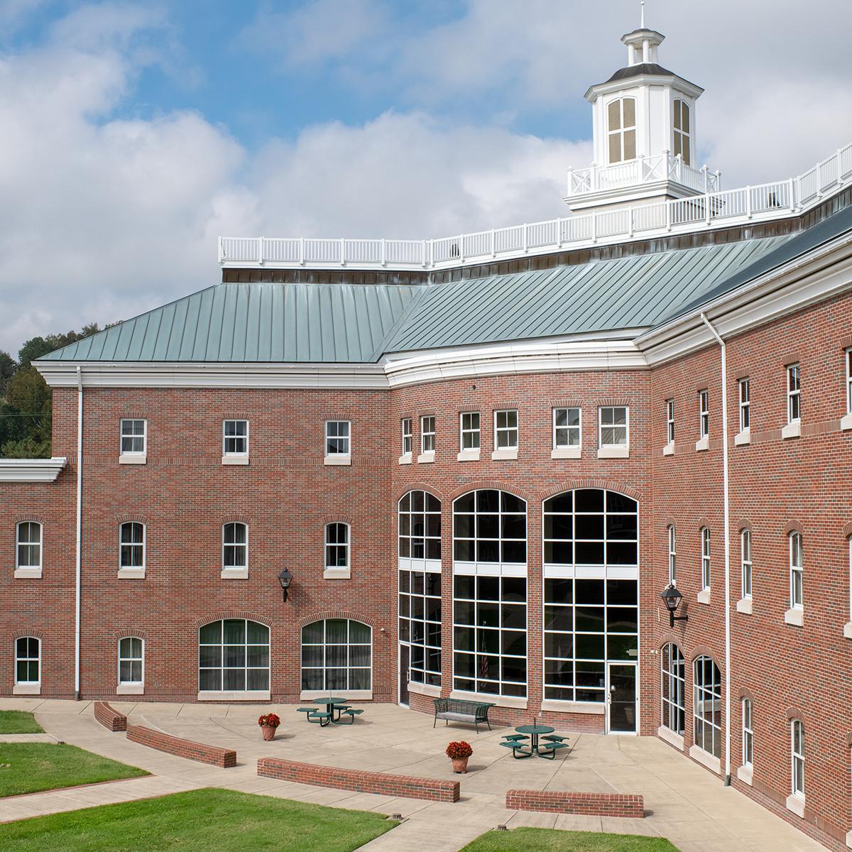 A three-story brick building with many windows. On the middle section, there is a large window that is two stories tall. It has a green tile rood and a white spire.