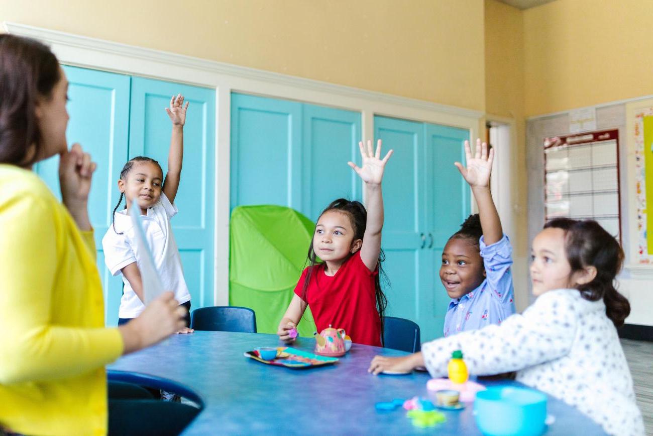 a teacher stands in front of young children with raised hands