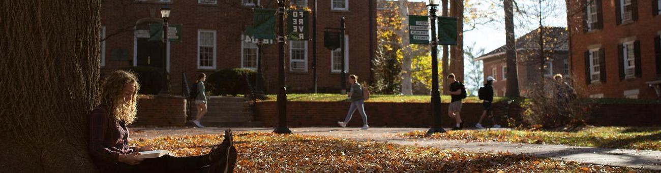 A student studies while sitting under a tree in front of Cutler Hall and McGuffey Hall as students walk by in the background.