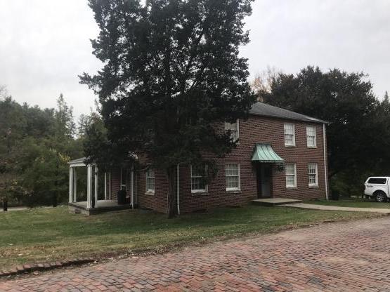 2-story rectangular brick building with front door looking out onto a brick street, with a side porch, and with a evergreen tree close to the corner of the building