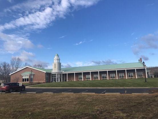 1-story brick building with white columns and clock tower, fronted by a lawn that gently slopes down to an asphalt parking lot